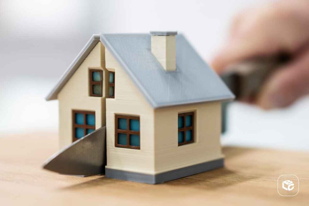 Miniatura de uma casa bege com telhado cinza sobre uma superfície de madeira. Uma lâmina metálica atravessa o centro da construção, criando a aparência de que a casa está sendo dividida em duas partes. Ao fundo, desfocado, aparece uma mão segurando o cabo da lâmina. A imagem simboliza divisão de patrimônio, partilha de bens, separação ou disputa imobiliária.