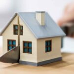 Miniatura de uma casa bege com telhado cinza sobre uma superfície de madeira. Uma lâmina metálica atravessa o centro da construção, criando a aparência de que a casa está sendo dividida em duas partes. Ao fundo, desfocado, aparece uma mão segurando o cabo da lâmina. A imagem simboliza divisão de patrimônio, partilha de bens, separação ou disputa imobiliária.